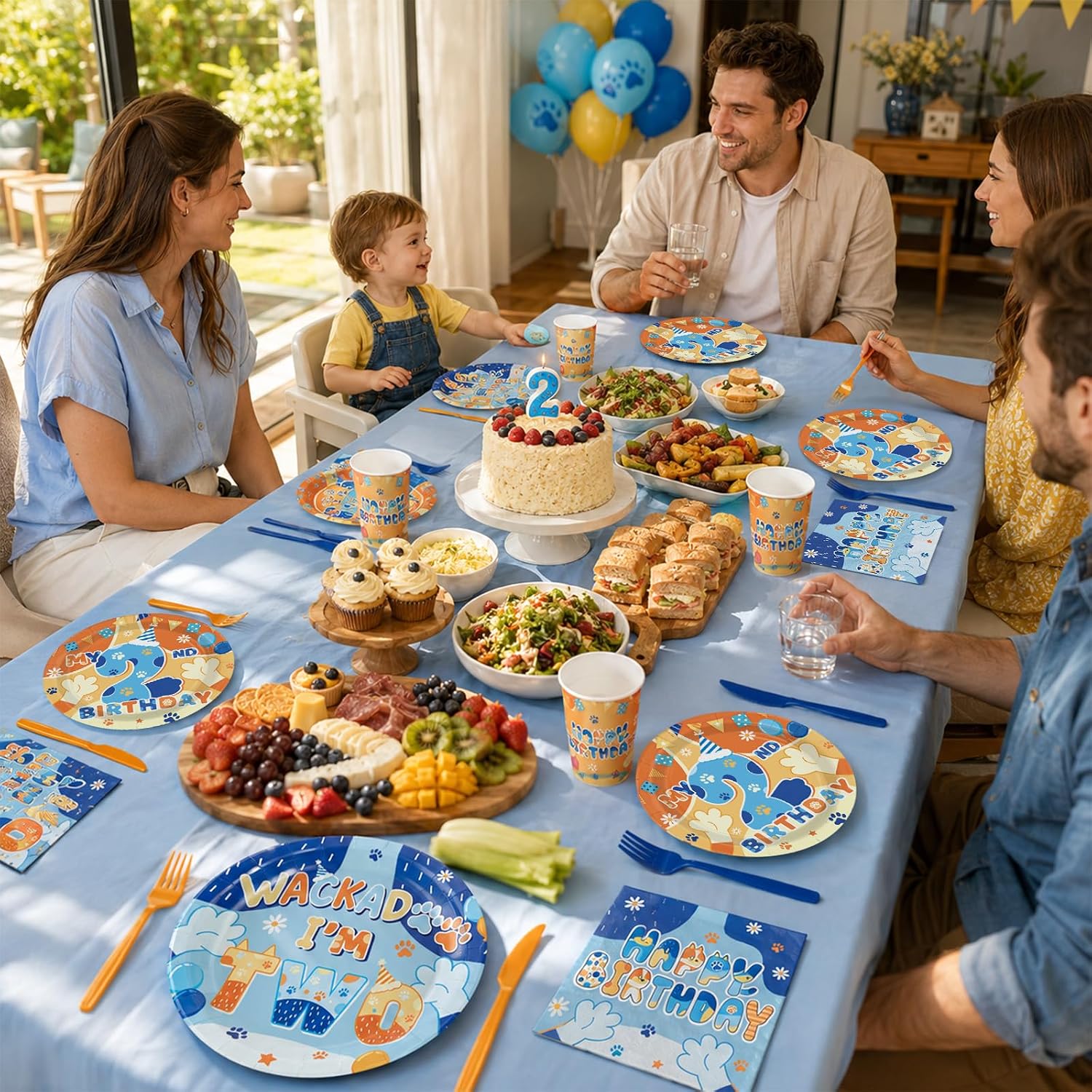 Family celebrating a 2nd birthday with Grencian blue dog tableware set featuring WACKADOO I am two plates, orange cups, blue napkins, blue and orange cutlery, balloon garland and birthday cake with number 2 candle